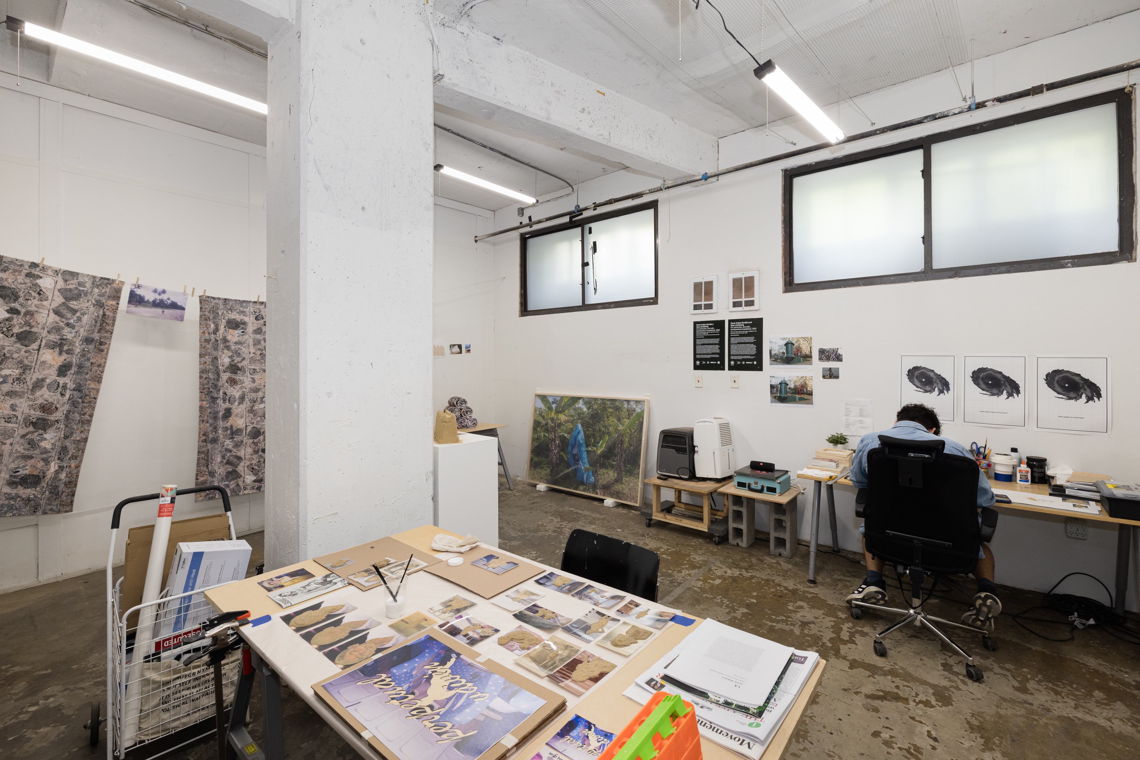 An artist studio with art and paper on a table and an artist working in the corner at a desk.