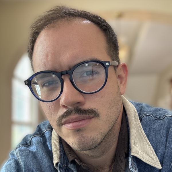 Photo of a man with a mustache wearing glasses and a denim jacket.